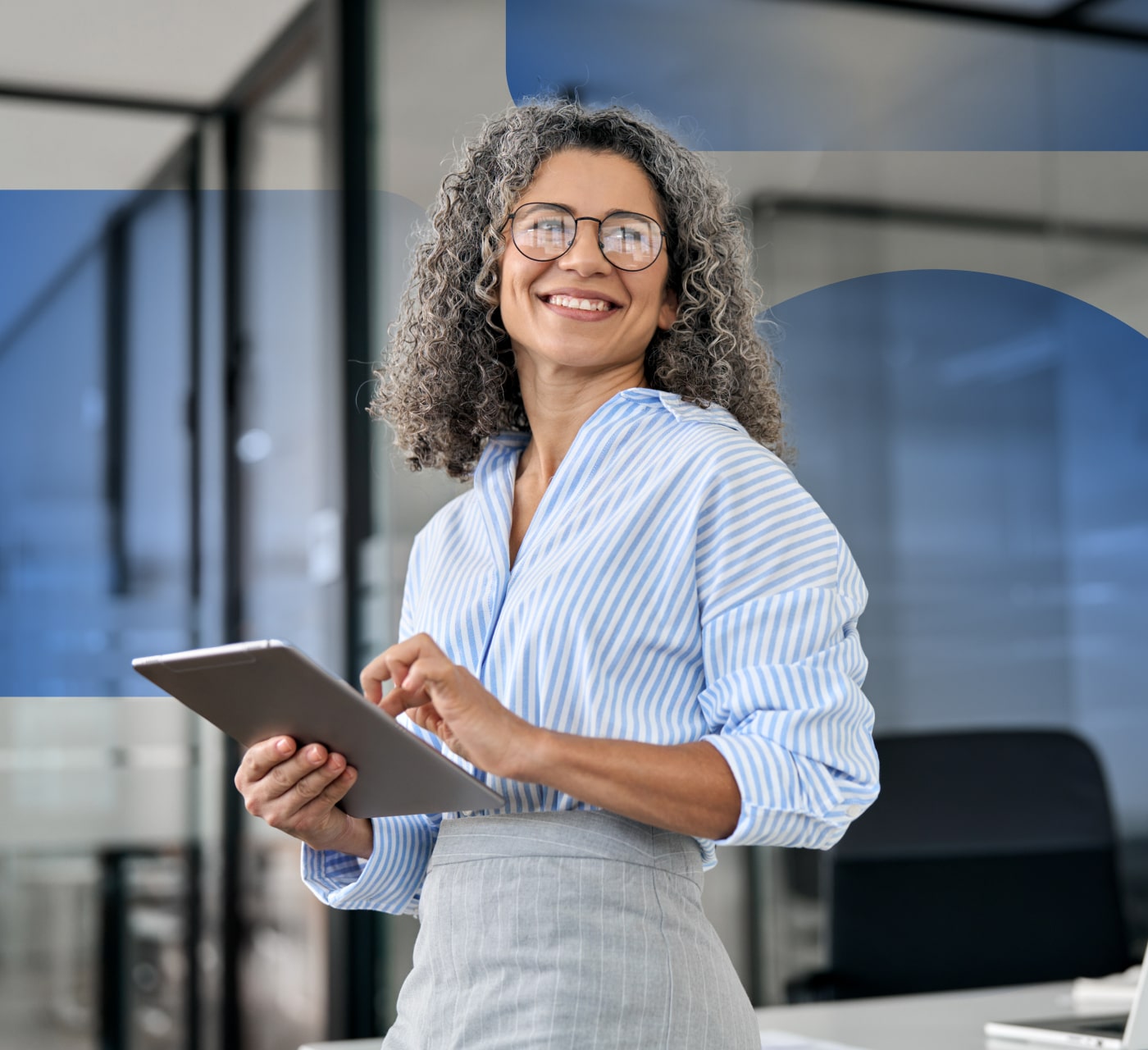 Woman in an office setting wearing a light blue shirt and grey skirt has a tablet in her hands and is smiling at the camera. She has grey curly hair and glasses.