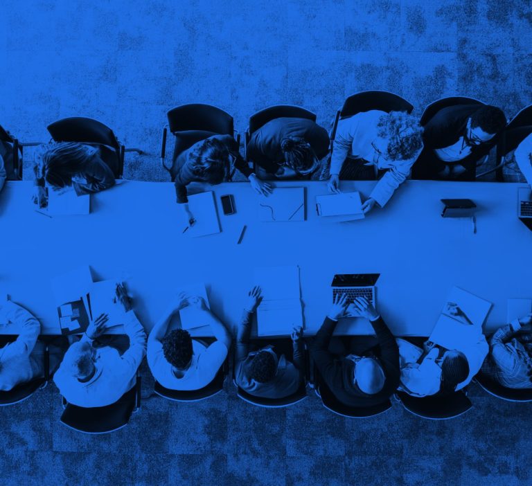 A group of 10 people sitting around a board table discussing complex project workflows. With a blue overlay on the image