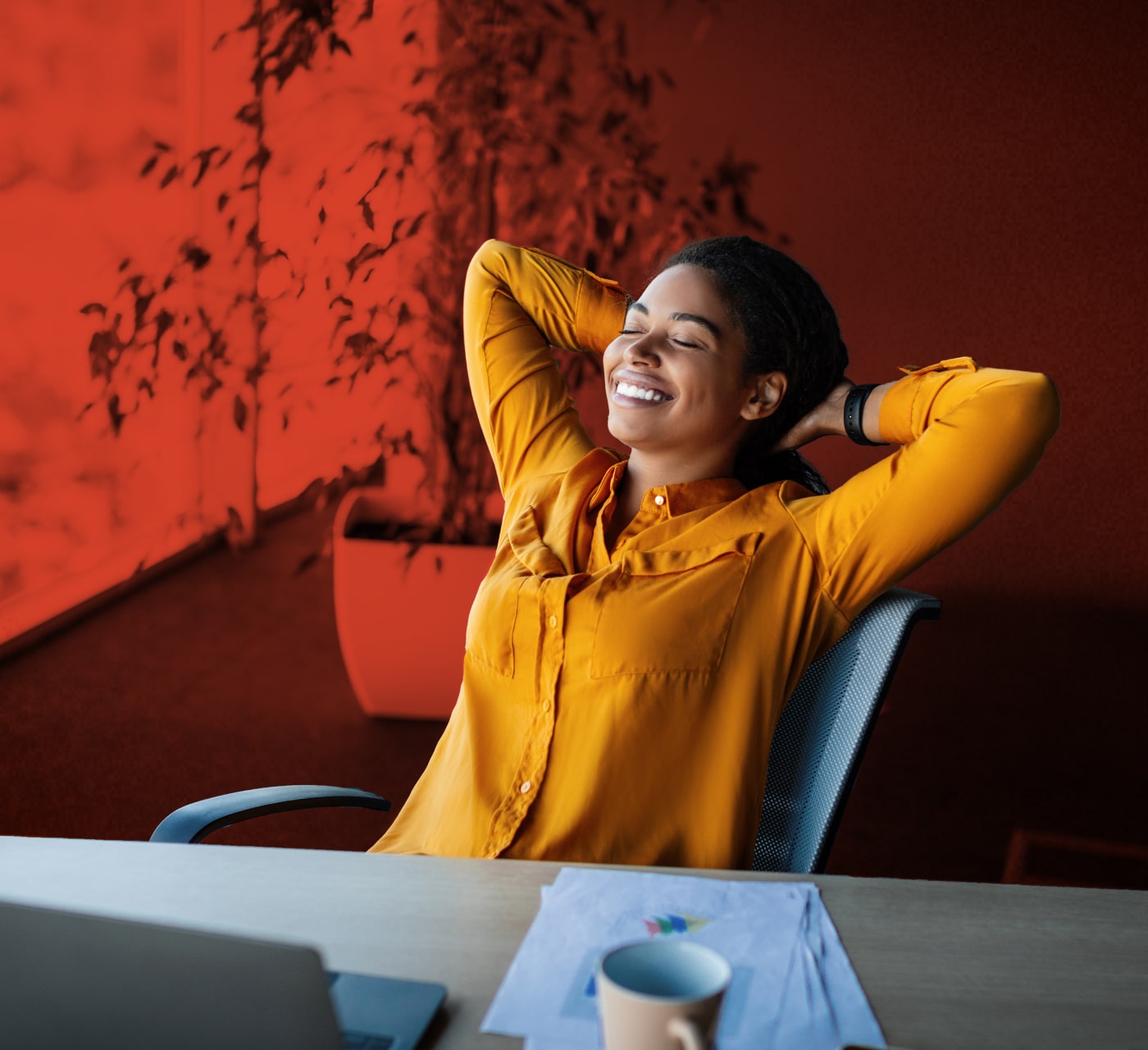 Business woman in an orange shirt leaning back in her desk chair with her arms behind her head with a laptop.