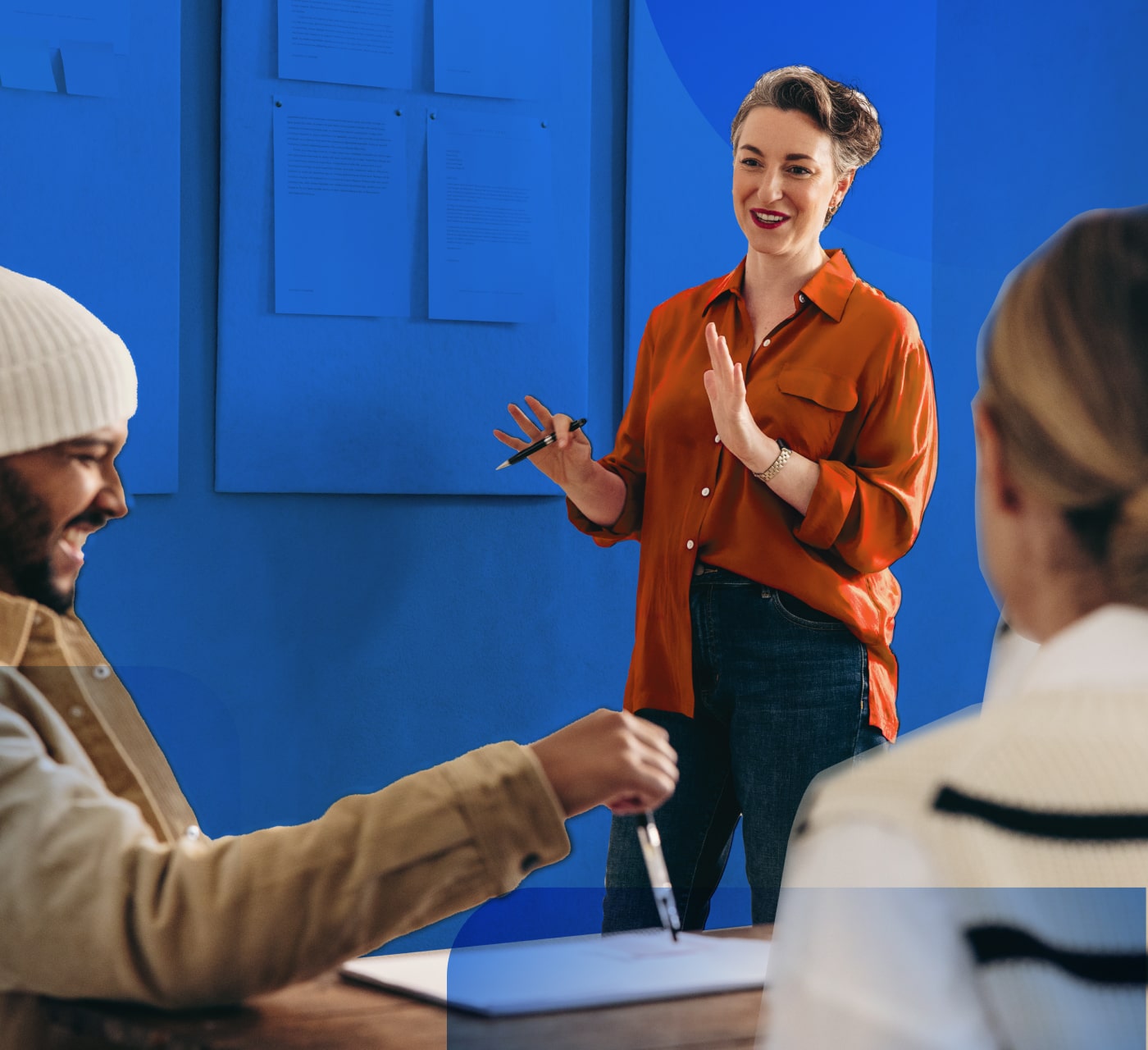 Woman with orange shirt while holding a pen presenting to two people on a blue background