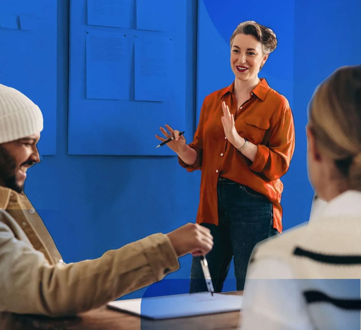 Woman with orange shirt while holding a pen presenting to two people on a blue background