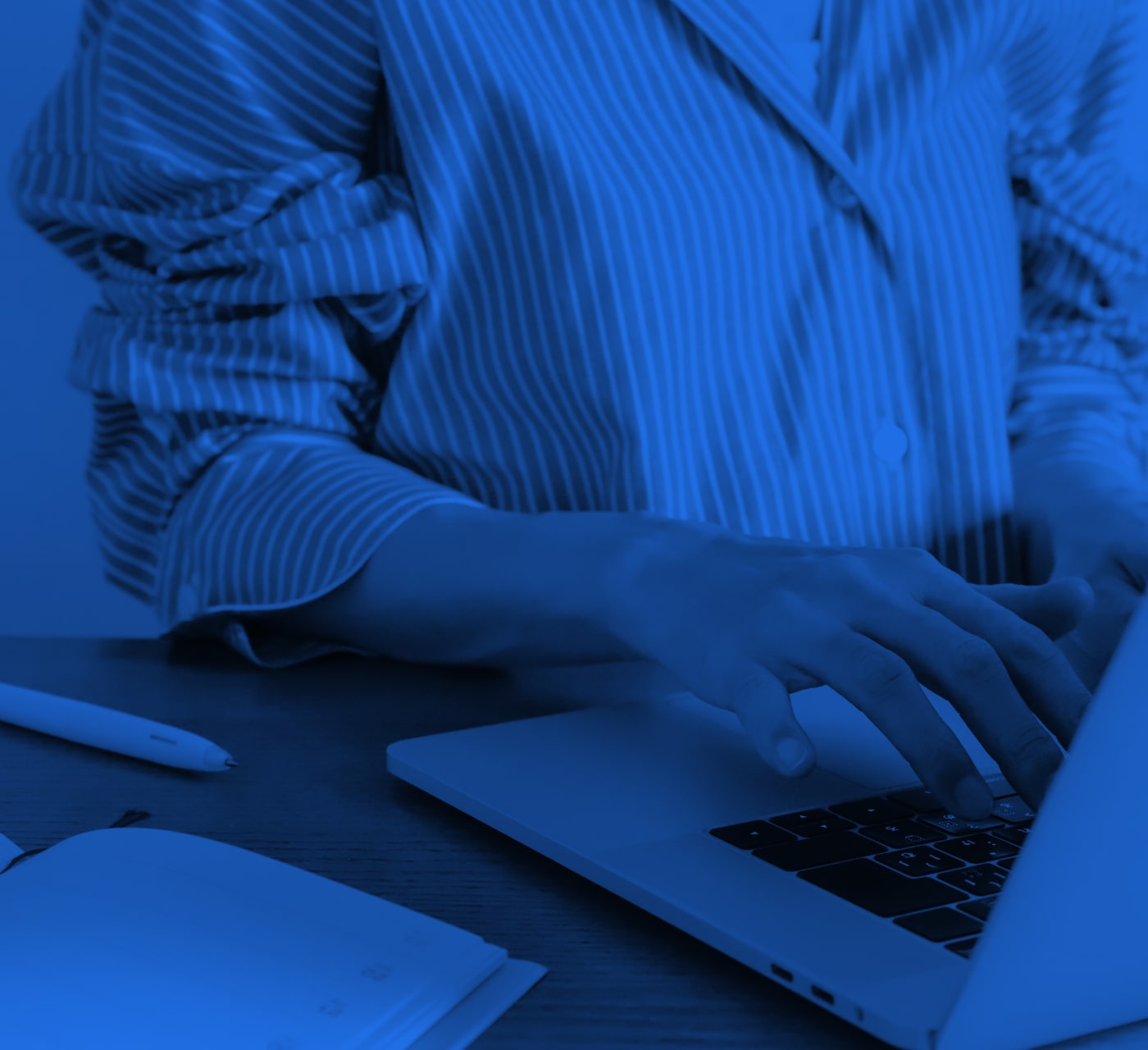 a person in a white shirt typing on a laptop keyboard with an open notebook beside them. Set to a blue background
