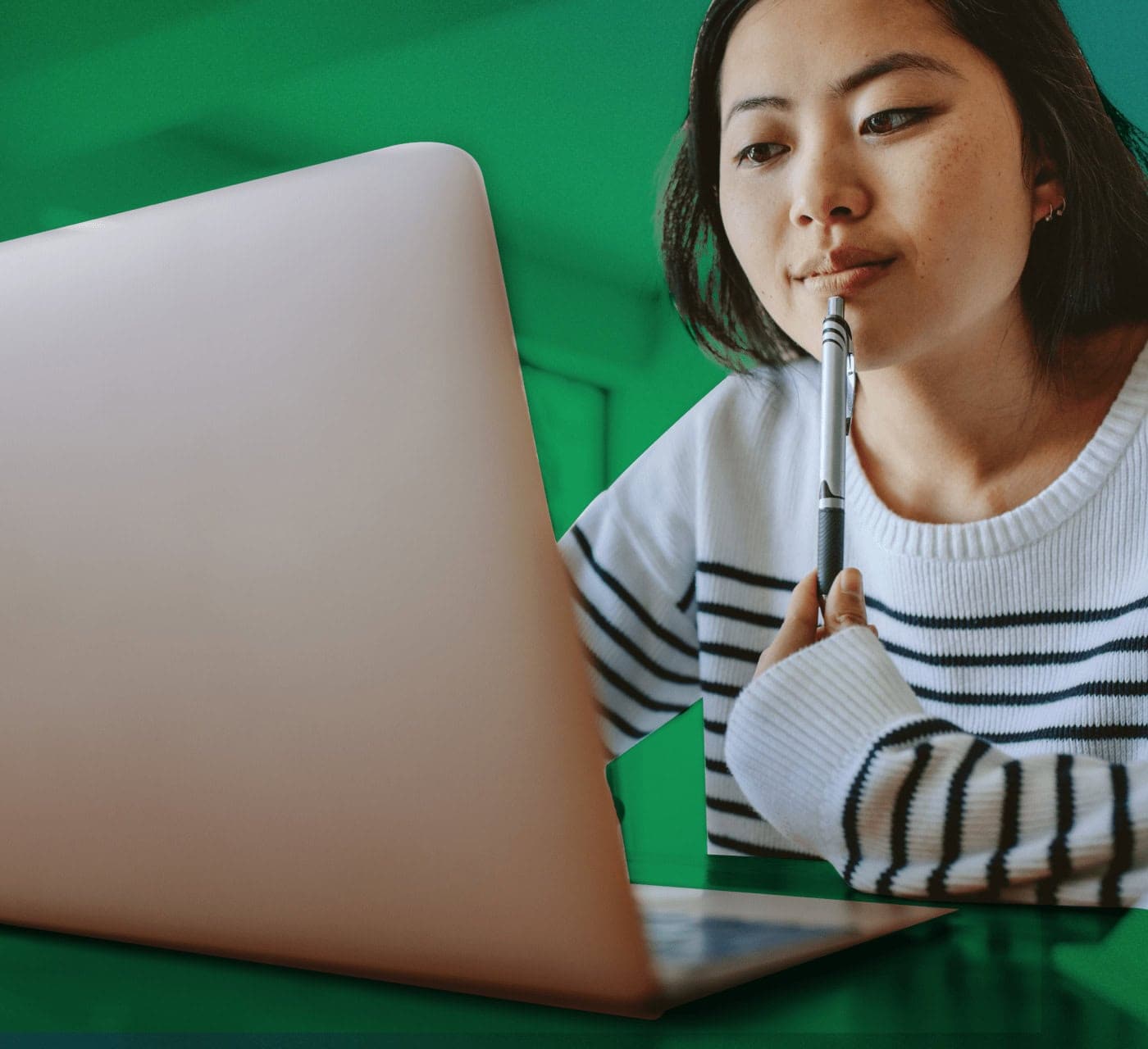 asian female with a pen in her hand smiling and looking at her laptop on a green background