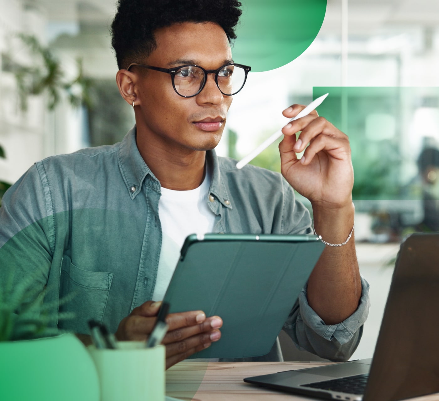 man in glasses and denim shirt with a pen and tablet in hand considering something he's writing. There is a laptop on the desk and the image is against an office background with green elements overlayed.