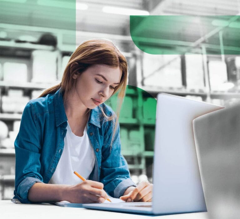 Woman in a denim shirt taking notes with a laptop open in front of her
